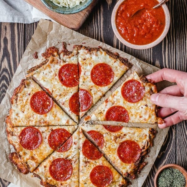 A hand grabs a slice of pepperoni pizza on parchment paper, with sauce and cheese nearby.