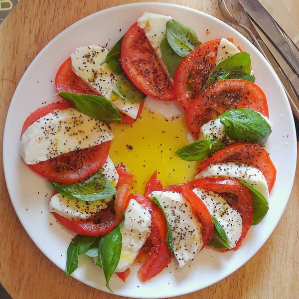 Caprese salad with sliced tomatoes, mozzarella, basil, olive oil, and pepper on a white plate.